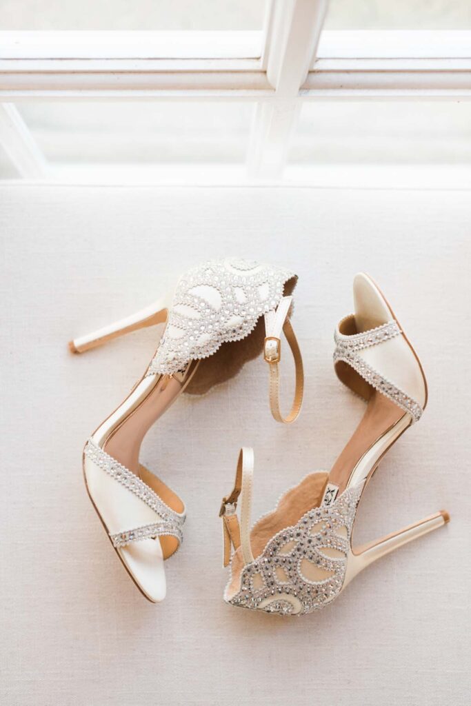 A pair of elegant high-heeled sandals with ankle straps, decorated with sparkling rhinestones and intricate beadwork, placed on a light-colored surface by a window. Copyright - Lisa Leanne Photography, California