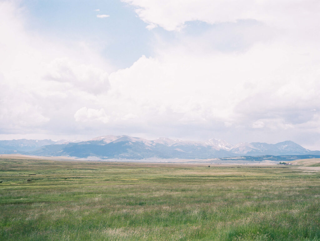 A wide, open grassland stretches into the distance with a range of low mountains under a partly cloudy sky, creating a serene and expansive landscape reminiscent of fine art film photography. Copyright - Lisa Leanne Photography, California