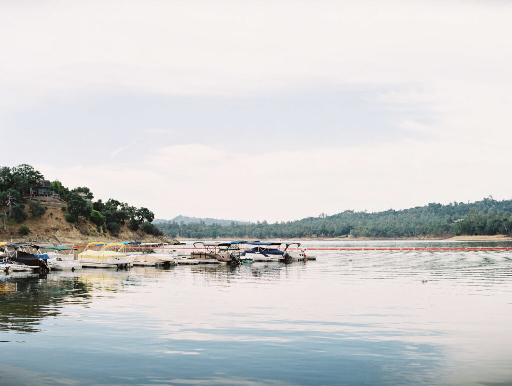 A row of small boats and pontoons are docked on a calm lake, with tree-covered hills in the background under a pale, cloudy sky. The scene, captured in fine art film photography, reflects softly on the tranquil water. Copyright - Lisa Leanne Photography, California