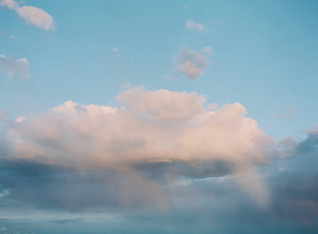 A soft, pastel-colored cloud floats in a blue sky, with subtle rays of sunlight shining through and faint streaks of rain beneath—evoking the dreamy atmosphere of fine art film photography. Copyright - Lisa Leanne Photography, California