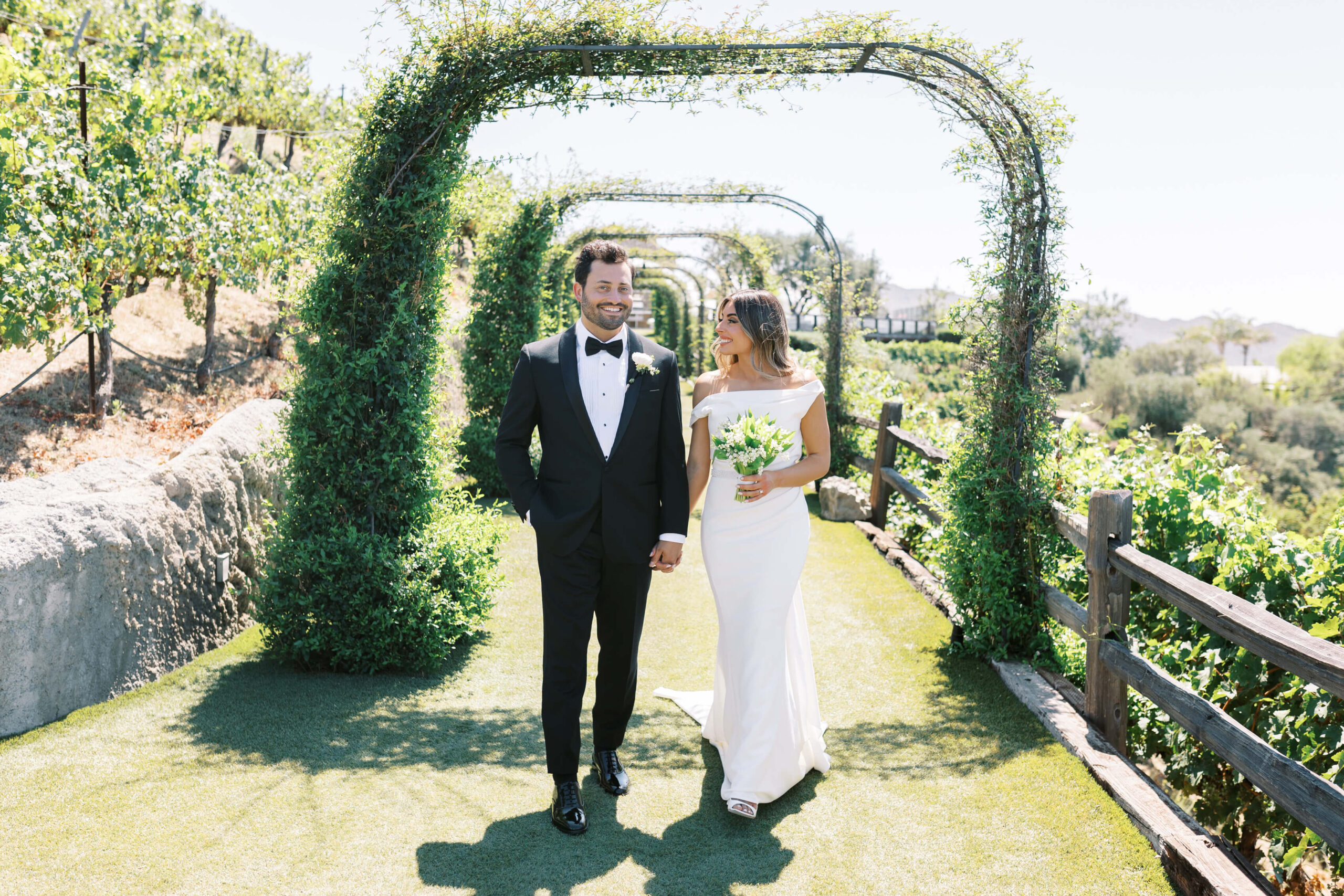 A bride and groom walk hand in hand outdoors under green vine-covered arches. The groom wears a black tuxedo, and the bride wears an off-the-shoulder white gown and holds a bouquet of flowers. It is a sunny day. Copyright - Lisa Leanne Photography, California