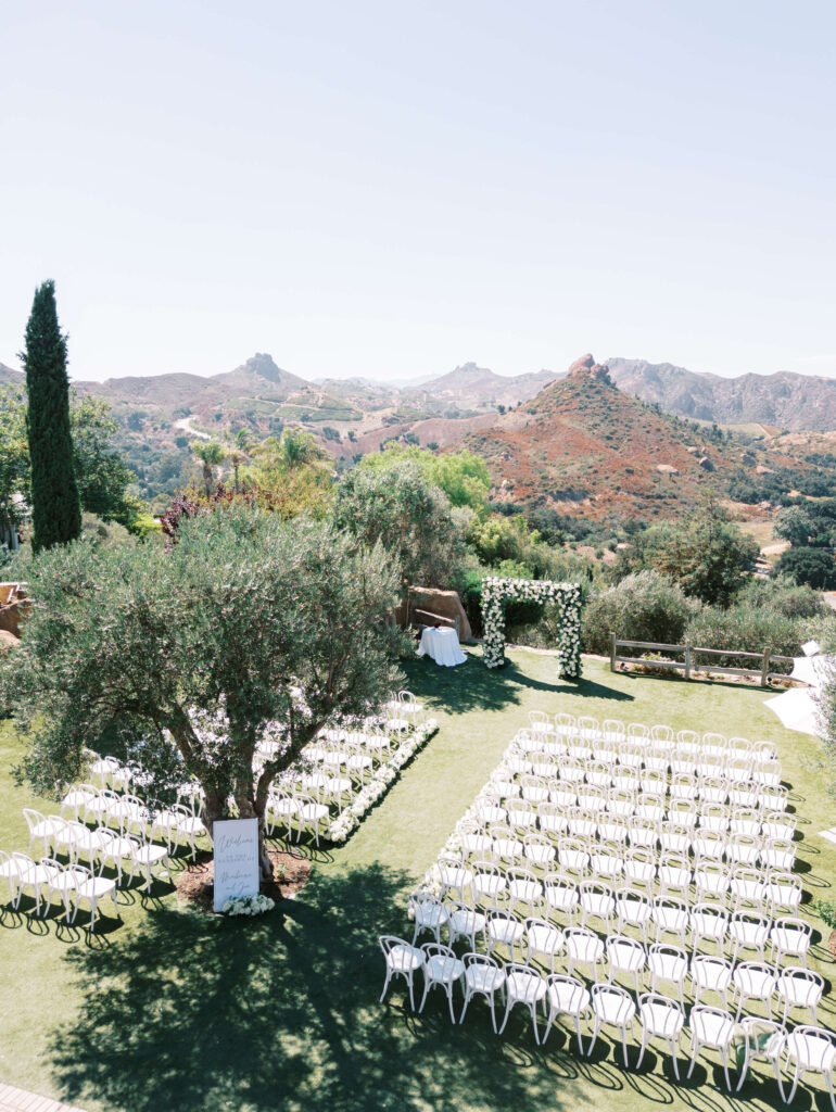 Outdoor wedding ceremony setup on a lawn with rows of white chairs, an aisle, lush greenery, an arch decorated with flowers, and scenic mountains in the background under a clear sky. Copyright - Lisa Leanne Photography, California