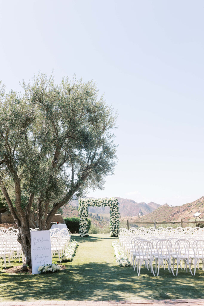 Outdoor wedding ceremony setup with white chairs arranged in rows facing a floral arch, set on green grass under a large tree, with mountains and clear sky in the background. A welcome sign stands to the left. Copyright - Lisa Leanne Photography, California