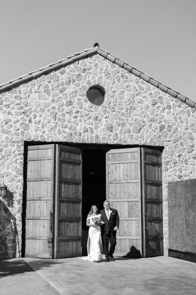 A bride in a white dress and a man in a suit stand arm-in-arm outside large wooden doors of a stone building, possibly at a wedding venue, on a sunny day. Copyright - Lisa Leanne Photography, California