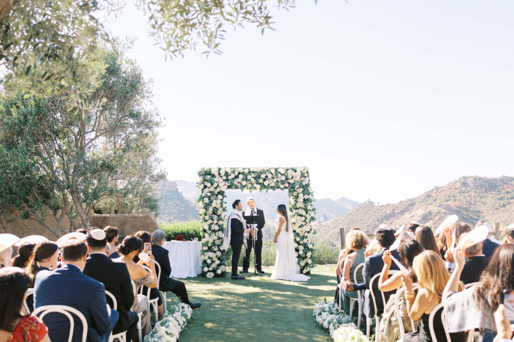 An outdoor wedding ceremony with guests seated on both sides of an aisle lined with flowers. A couple stands in front of a floral arch, exchanging vows with mountains visible in the background under a clear sky. Copyright - Lisa Leanne Photography, California