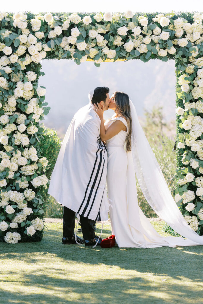 A bride and groom kiss under a wedding arch decorated with white flowers. The groom wears a white prayer shawl with black stripes; the bride wears a white dress and veil. It is an outdoor ceremony with greenery in the background. Copyright - Lisa Leanne Photography, California