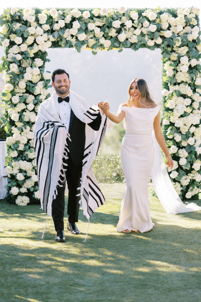 A joyful bride and groom walk hand in hand outdoors under a floral arch, with the groom wearing a black tuxedo and a tallit, and the bride in a white gown, both smiling. Copyright - Lisa Leanne Photography, California