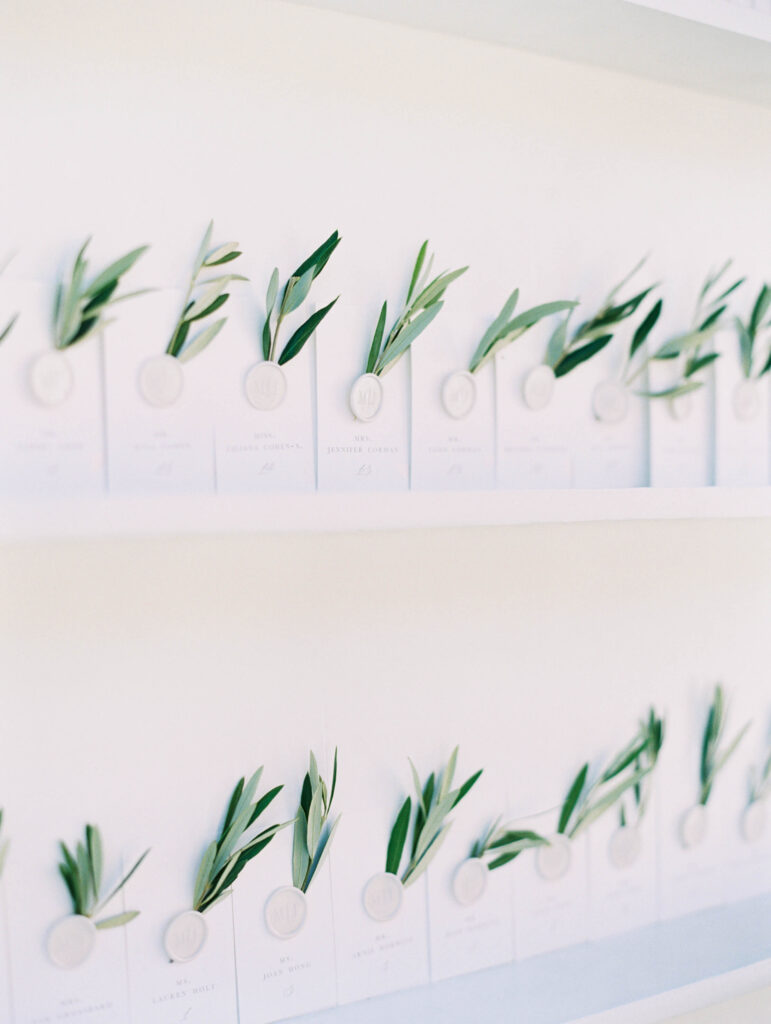 Rows of white cards, each decorated with a wax seal and a small sprig of green leaves, are neatly arranged on white shelves against a light background. Copyright - Lisa Leanne Photography, California