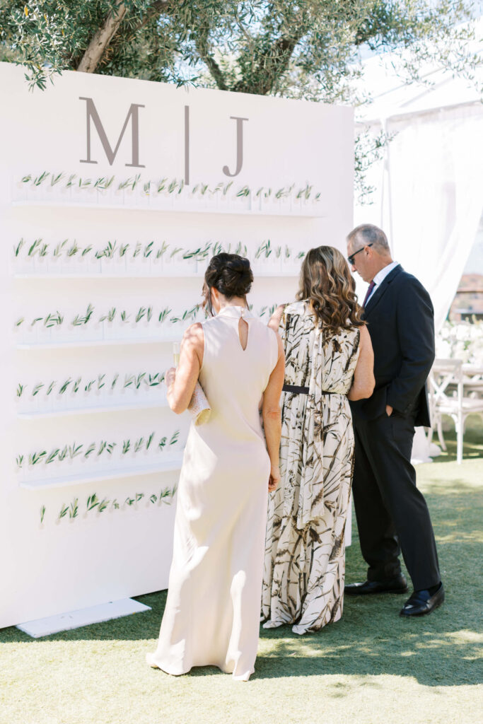 Three people in formal attire look at a white display wall with greenery and cards under the large initials “M | J” at an outdoor event, possibly a wedding. Bright, sunny setting with trees and chairs in the background. Copyright - Lisa Leanne Photography, California