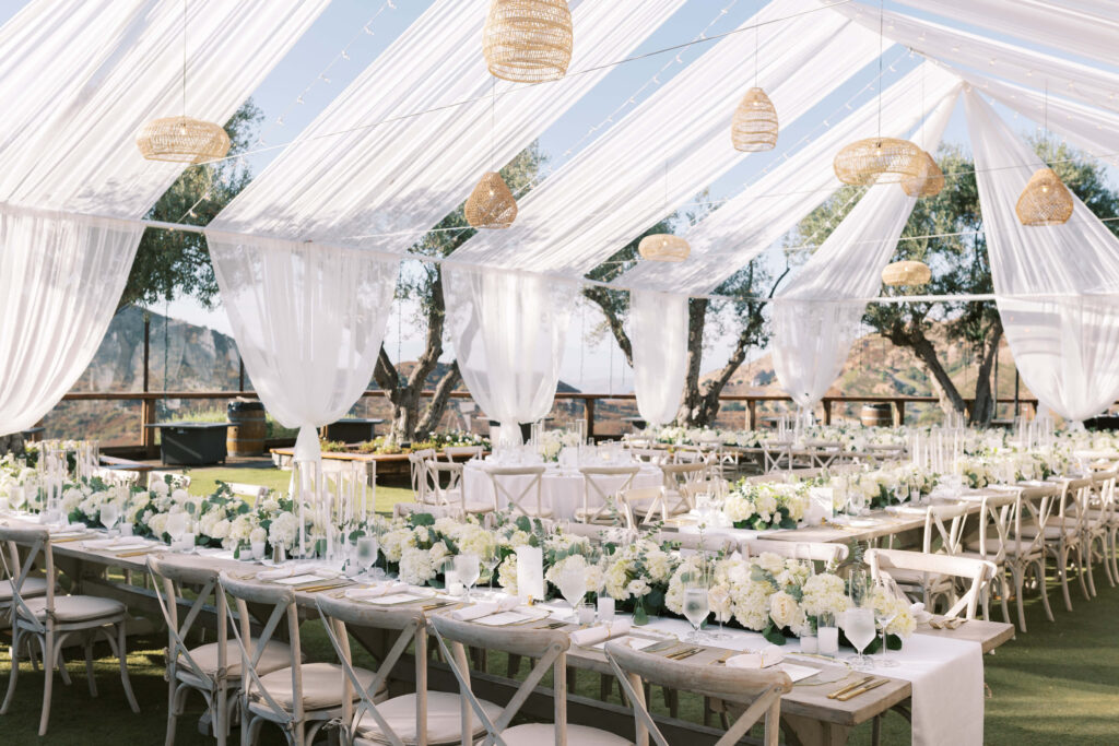 Elegant outdoor wedding reception under a clear tent with white drapes, hanging woven lanterns, and long tables decorated with white flowers, greenery, and gold accents set for guests. Copyright - Lisa Leanne Photography, California