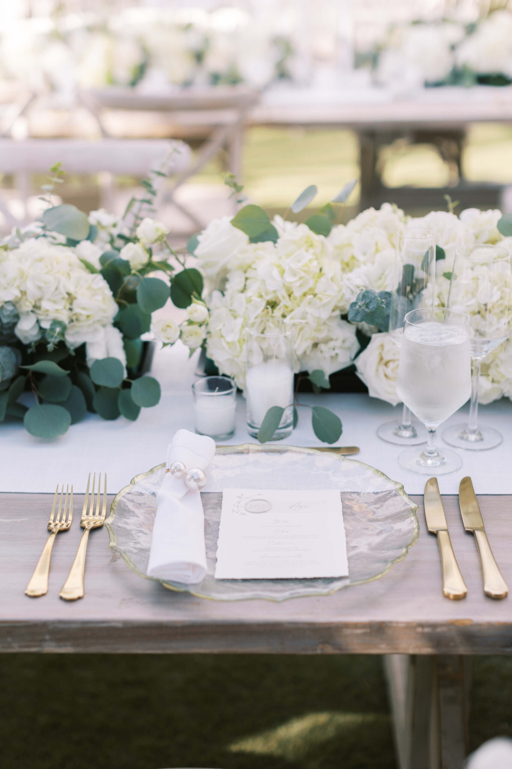 Elegant wedding table setting with gold cutlery, a clear glass plate, a rolled napkin, and a printed menu. The table is decorated with white flowers and greenery, and has a glass of water and wine glasses. Copyright - Lisa Leanne Photography, California