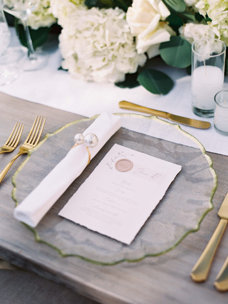 A close-up of an elegant table setting with a gold-rimmed glass plate, gold flatware, a white napkin with a pearl napkin ring, a printed menu, and white flowers in the background. Copyright - Lisa Leanne Photography, California