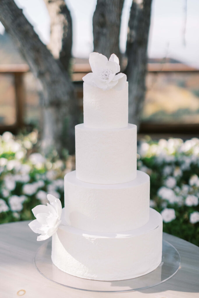A four-tiered white wedding cake decorated with two large white sugar flowers, one on top and one on the side, displayed outdoors with trees and white flowers in the background. Copyright - Lisa Leanne Photography, California