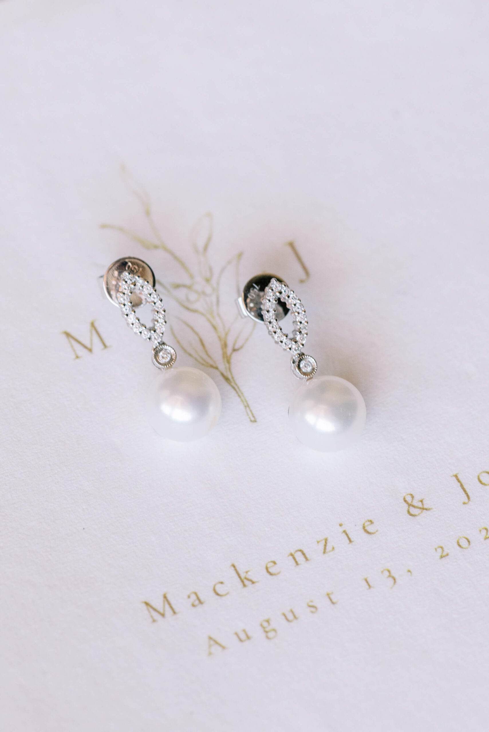 A pair of elegant pearl drop earrings with diamond accents rests on white stationery featuring gold text and a botanical illustration. Copyright - Lisa Leanne Photography, California