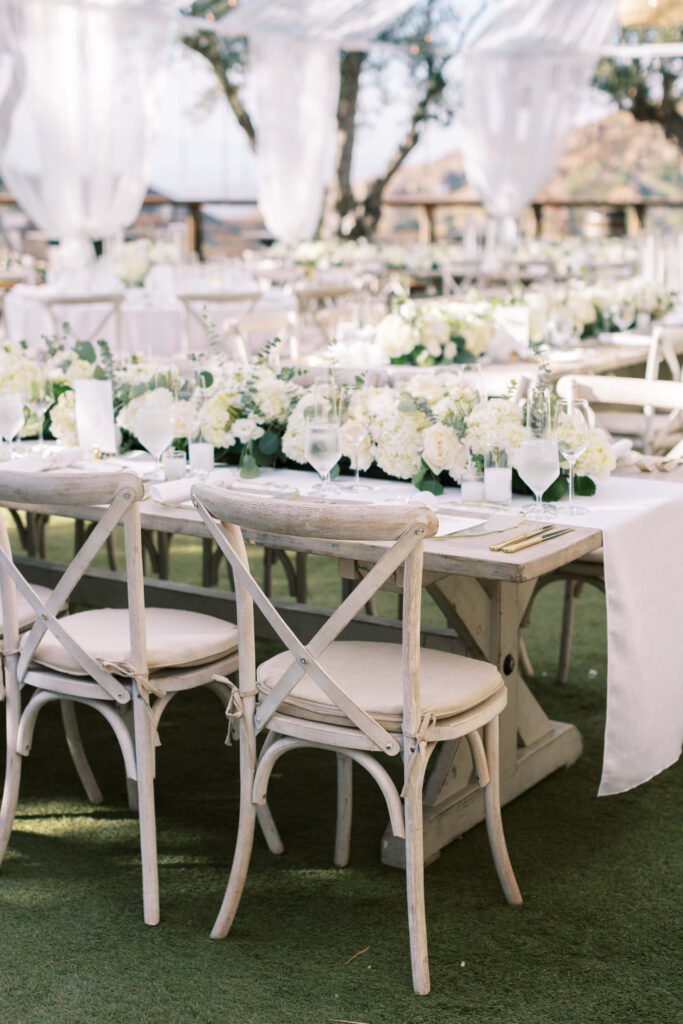 Elegant outdoor wedding reception setup with white wooden chairs, long tables covered in white tablecloths, and floral centerpieces featuring white flowers and greenery. Sheer drapes hang above, creating a soft, romantic atmosphere. Copyright - Lisa Leanne Photography, California
