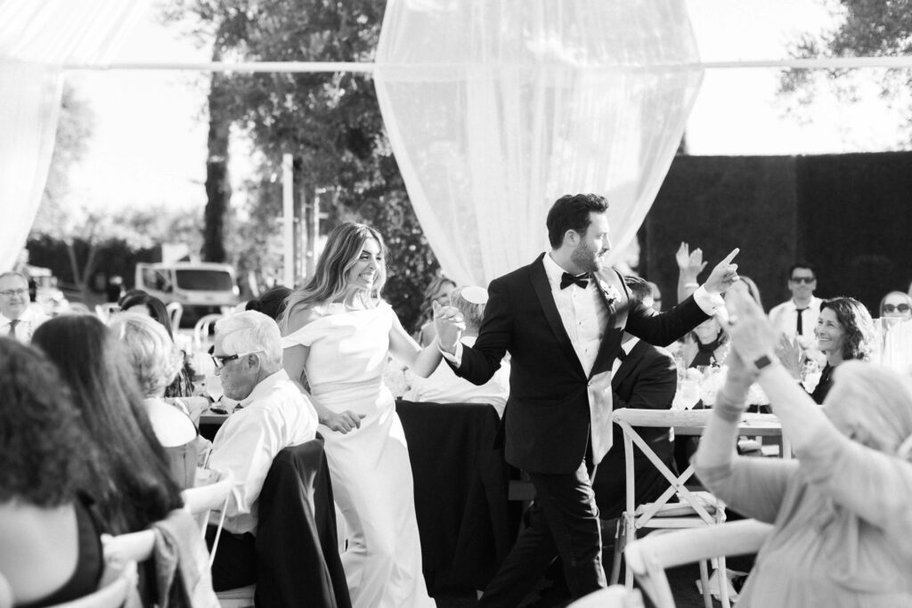 A bride and groom, smiling and holding hands, walk among seated wedding guests outdoors. Guests applaud and cheer as sunlight filters through sheer drapes. The scene is joyful and celebratory. Copyright - Lisa Leanne Photography, California