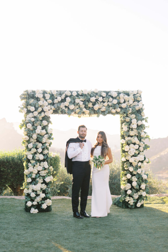 A bride in a white dress and a groom in a white shirt and black pants stand under a square floral arch made of white flowers and greenery, outdoors with mountains in the background. Copyright - Lisa Leanne Photography, California