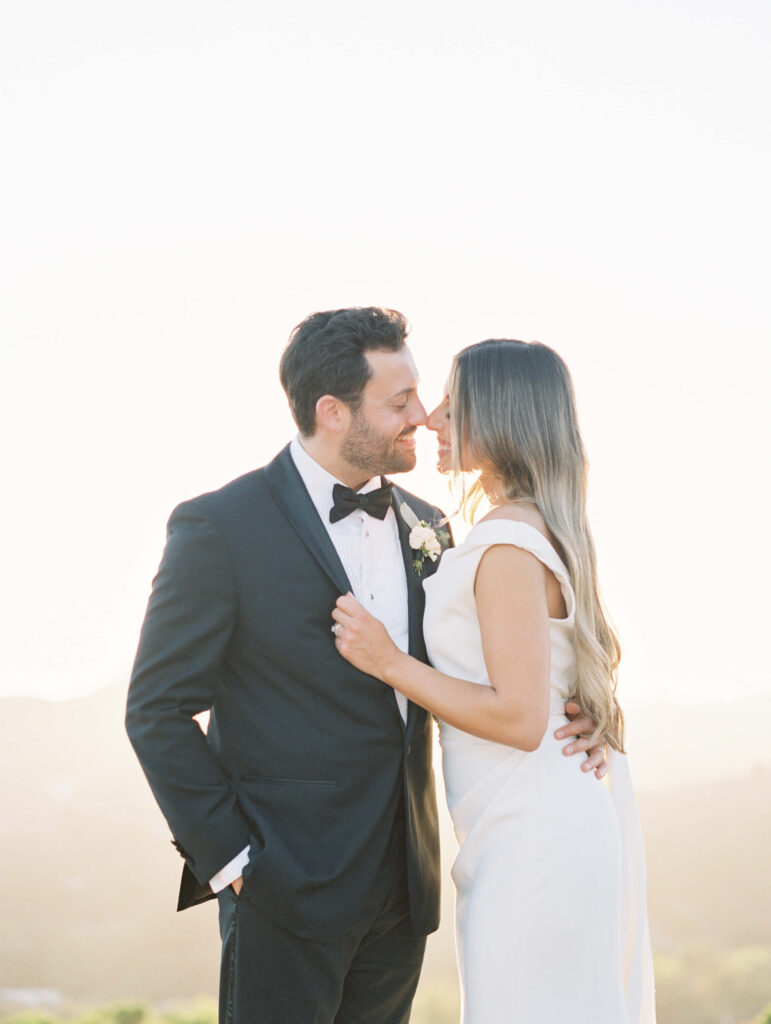 A bride in a white dress and a groom in a black tuxedo stand close, facing each other and about to kiss, with soft sunlight illuminating them from behind. Copyright - Lisa Leanne Photography, California