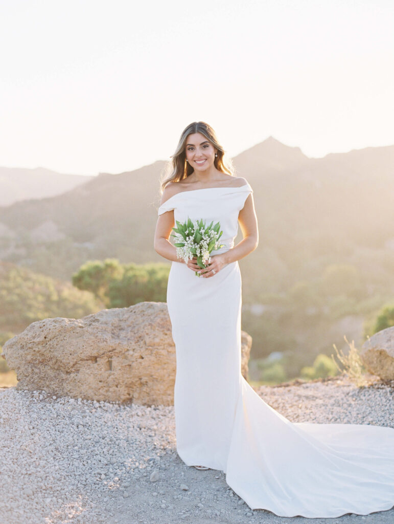A bride in an off-the-shoulder white gown holds a small bouquet of flowers, standing on rocky ground with sunlit hills and greenery in the background. Copyright - Lisa Leanne Photography, California