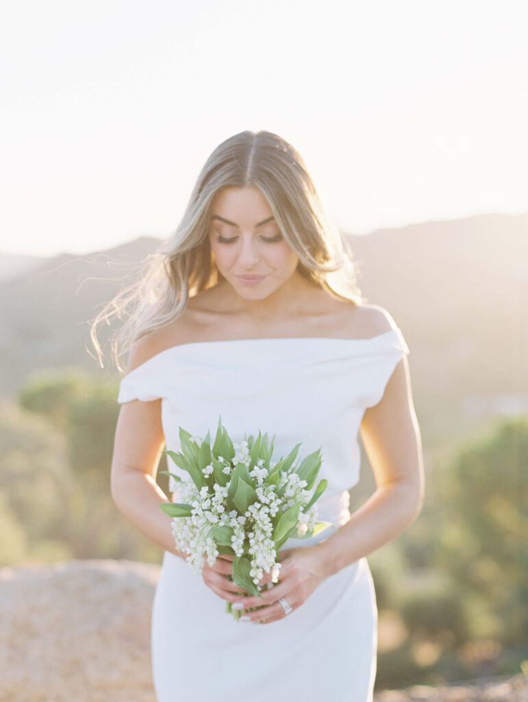 A woman in an off-the-shoulder white dress stands outdoors, holding a bouquet of lily of the valley flowers. The background is softly lit with natural sunlight and blurred greenery. Copyright - Lisa Leanne Photography, California