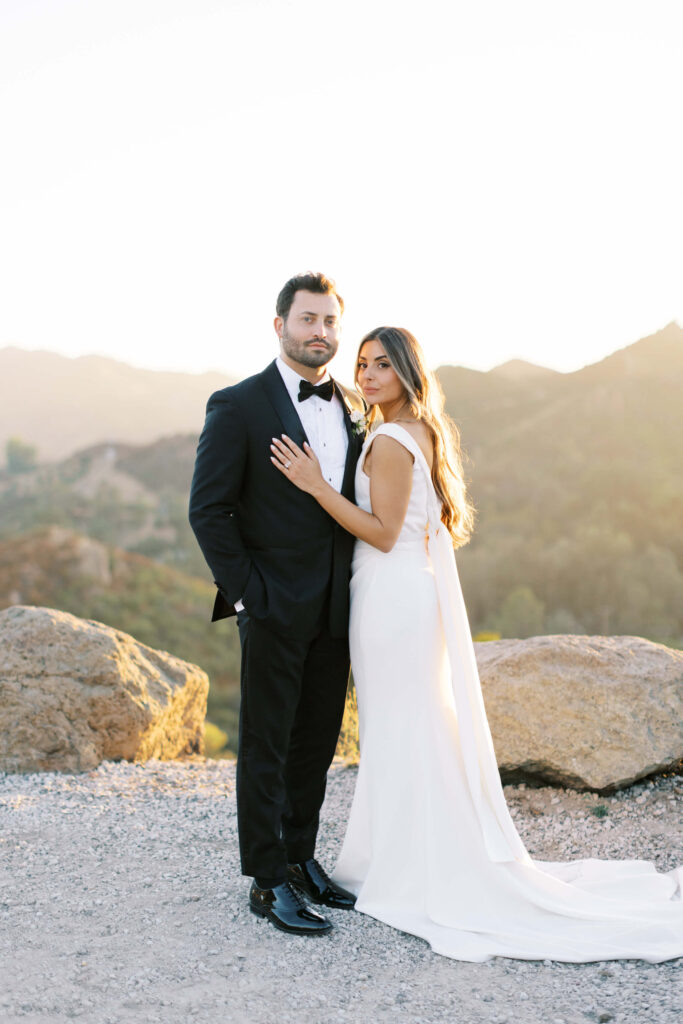 A bride in a white gown and groom in a black tuxedo pose together at golden hour, with sunlit mountains and boulders in the background. Copyright - Lisa Leanne Photography, California