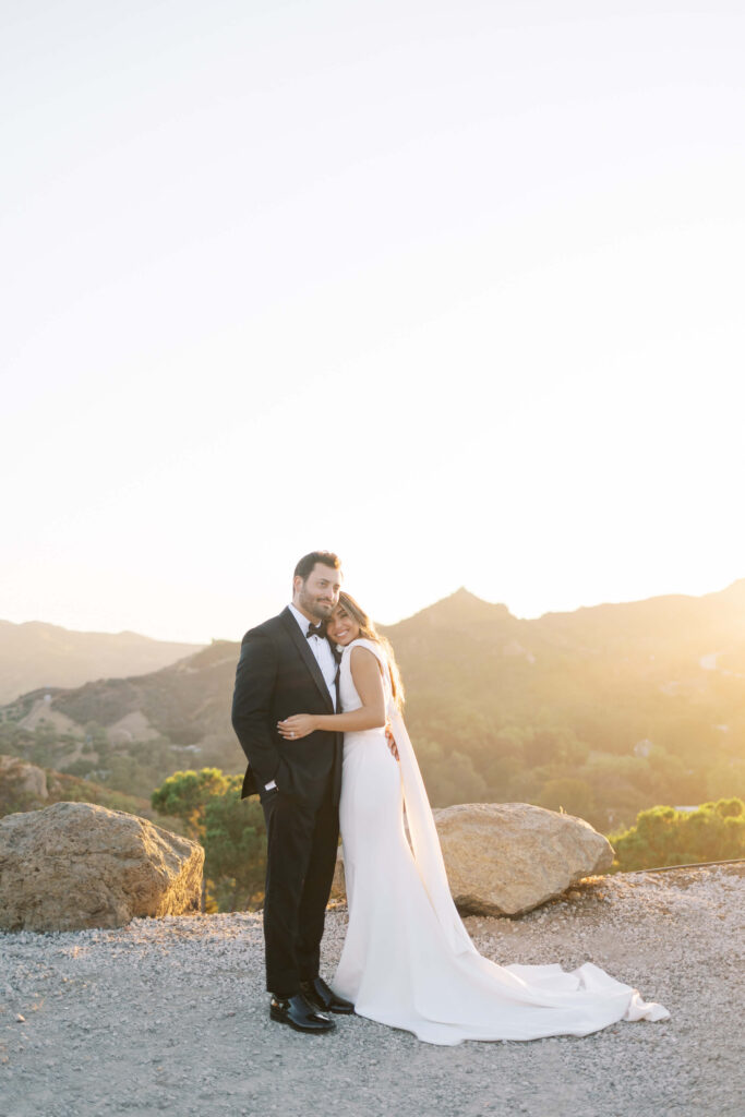 A bride in a white dress hugs and leans her head on a groom in a black suit. They stand outdoors at sunset with mountains in the background and large rocks nearby. Copyright - Lisa Leanne Photography, California
