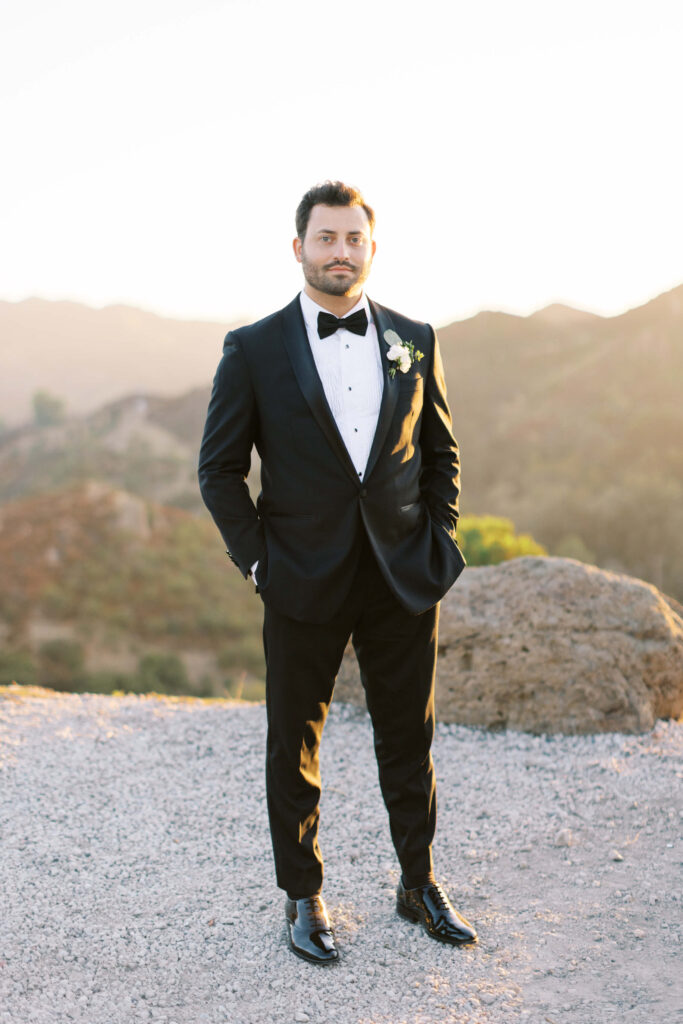 A man in a black tuxedo with a bow tie and boutonniere stands outdoors on a gravel path, with mountains and greenery in the background at sunset. Copyright - Lisa Leanne Photography, California