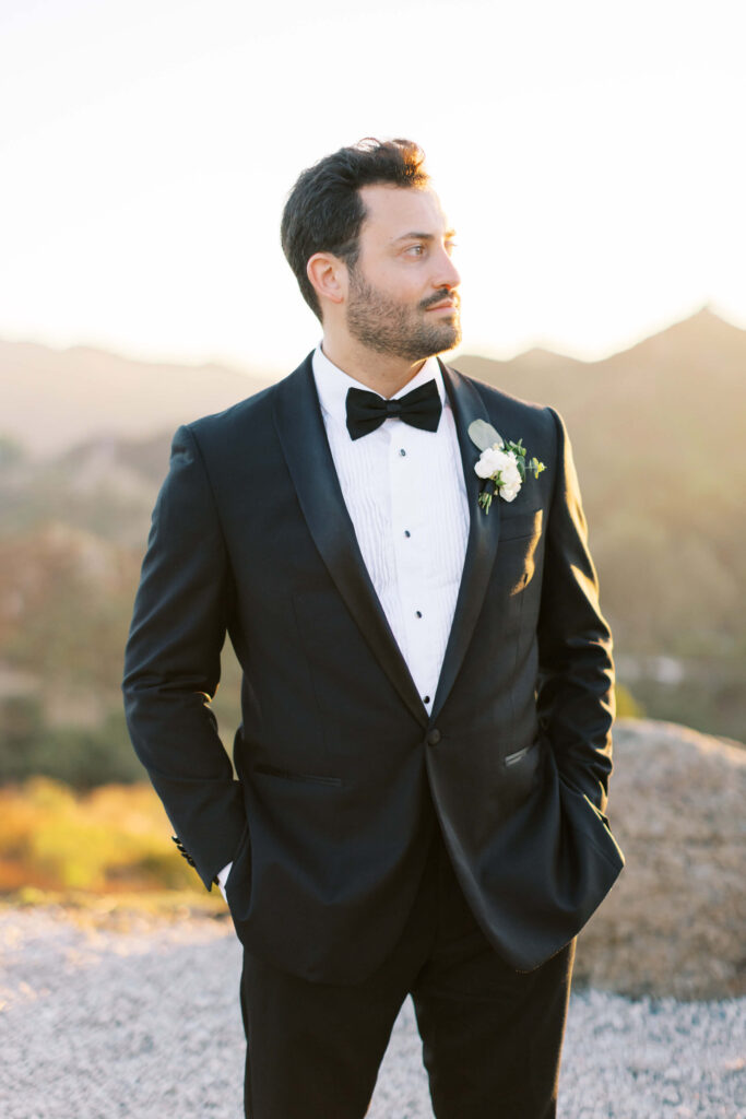 A man in a black tuxedo with a white shirt and black bow tie stands outdoors with hands in his pockets, looking to the side. He wears a boutonnière on his lapel, and the background is softly blurred with natural scenery. Copyright - Lisa Leanne Photography, California