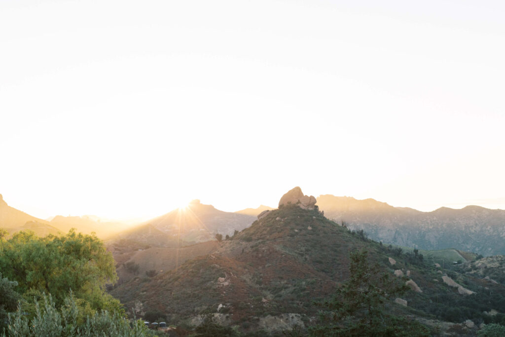 The sun rises over distant mountains, casting a soft golden light across rolling hills and rocky peaks, with scattered trees and greenery in the foreground under a clear sky. Copyright - Lisa Leanne Photography, California