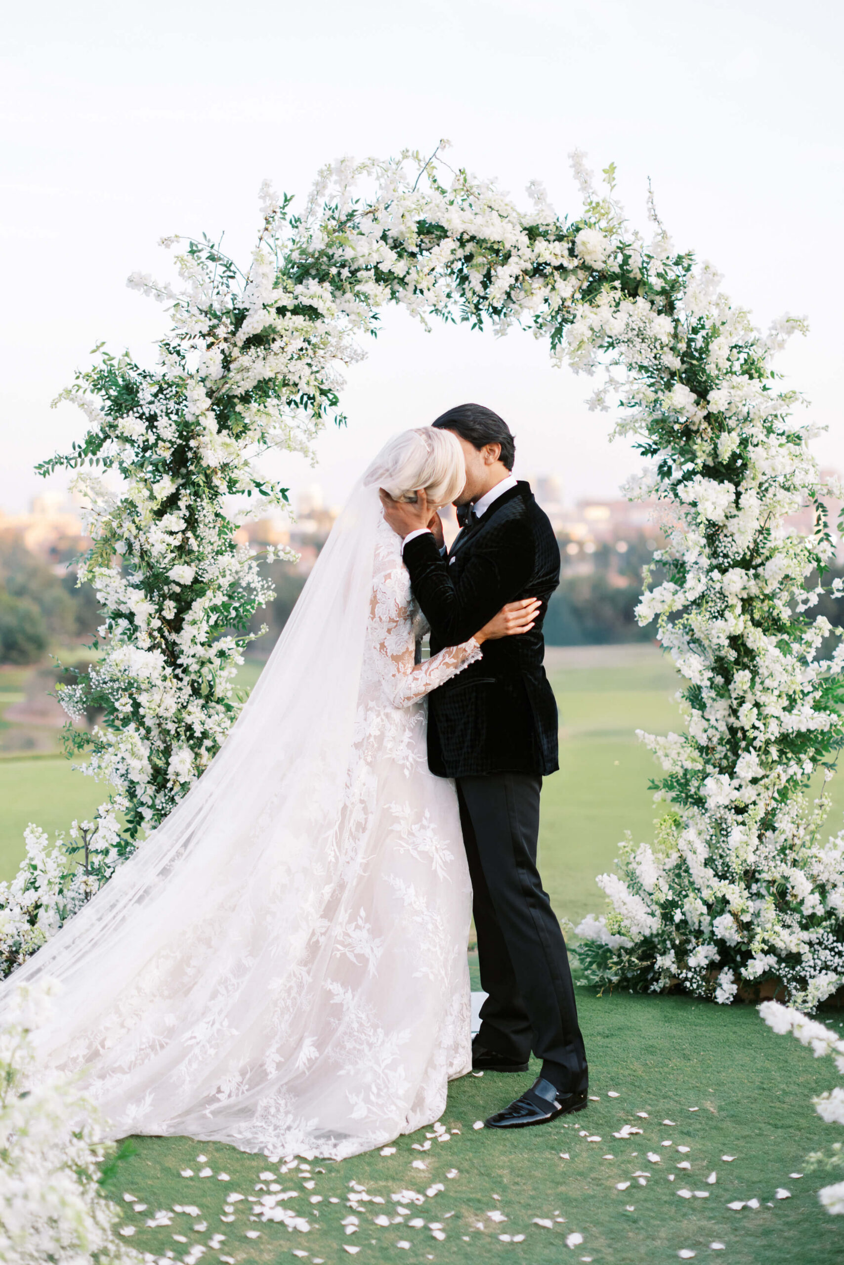 A bride and groom embrace and kiss under a large, elegant floral arch outdoors at their Bel-Air Country Club wedding. The bride wears a long white gown and veil, while the groom is in a black suit. White flower petals cover the grass around them. Copyright - Lisa Leanne Photography, California