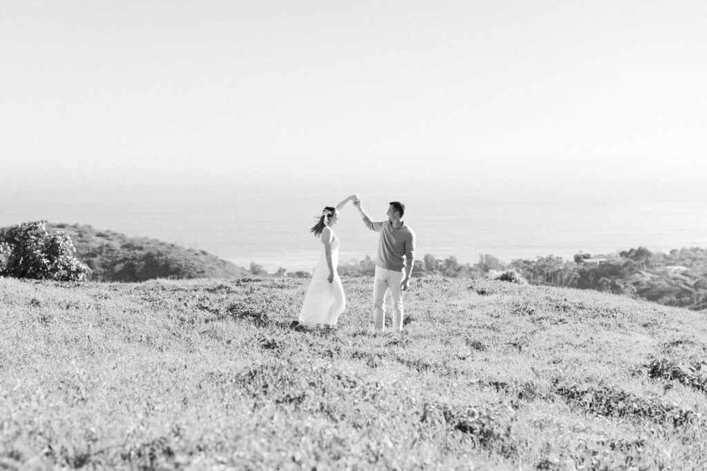 A couple dances together in a sunlit, grassy field overlooking the ocean. The woman wears a white dress, and the man lifts her hand as they smile, surrounded by nature under a clear sky. Copyright - Lisa Leanne Photography, California