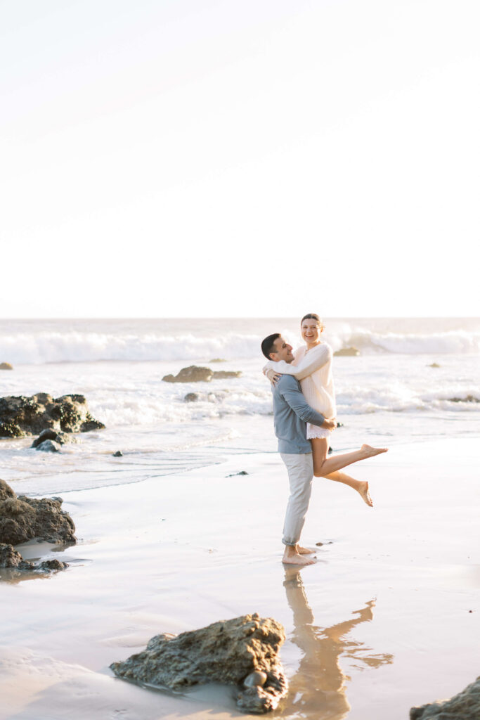 A man in light-colored clothes lifts a smiling woman, both barefoot, on a sandy beach near rocks with gentle ocean waves in the background under a bright sky. Copyright - Lisa Leanne Photography, California