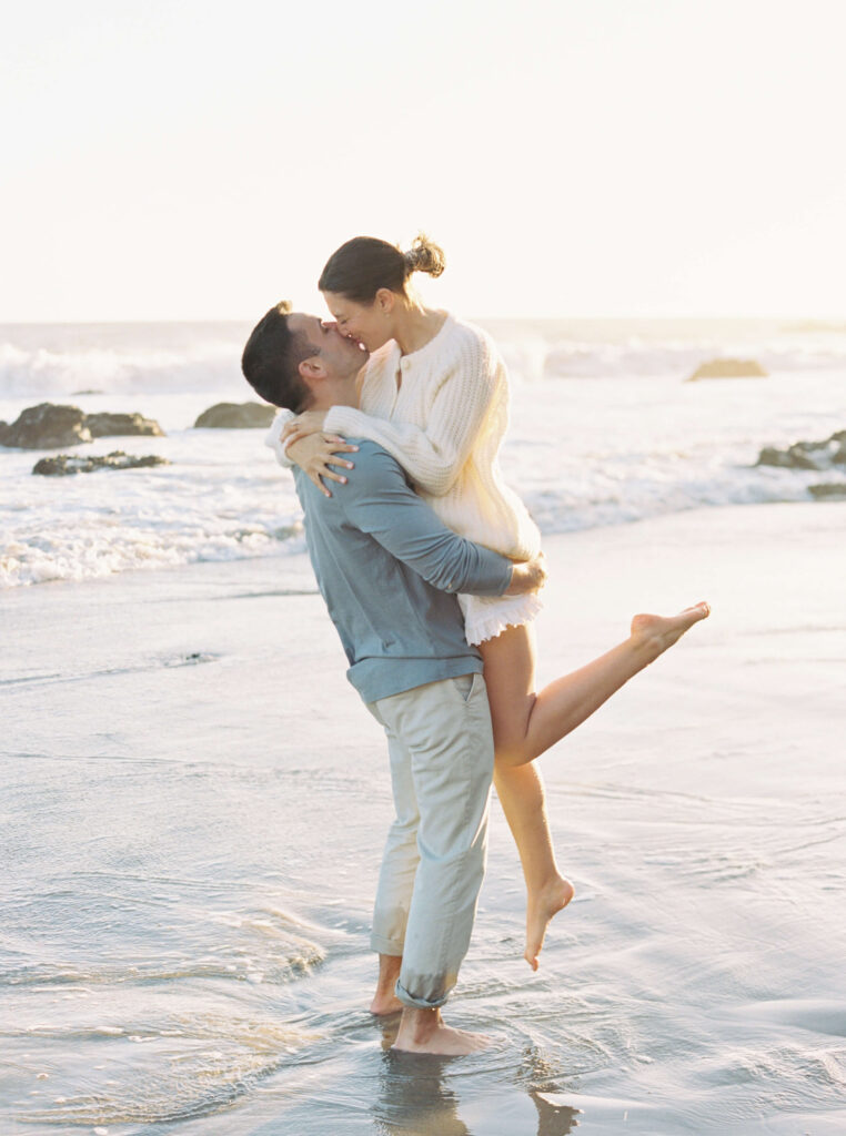 A man stands barefoot in shallow ocean water, lifting a smiling woman in a white sweater and dress. They embrace and laugh together, with gentle waves and a bright, sunlit sky in the background. Copyright - Lisa Leanne Photography, California