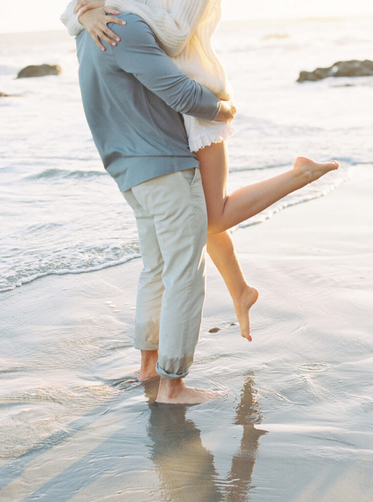 A couple stands barefoot on the beach at the water&rsquo;s edge; one person lifts the other in an embrace, with the lifted person's legs bent and one foot off the ground, while soft sunlight illuminates the scene. Copyright - Lisa Leanne Photography, California