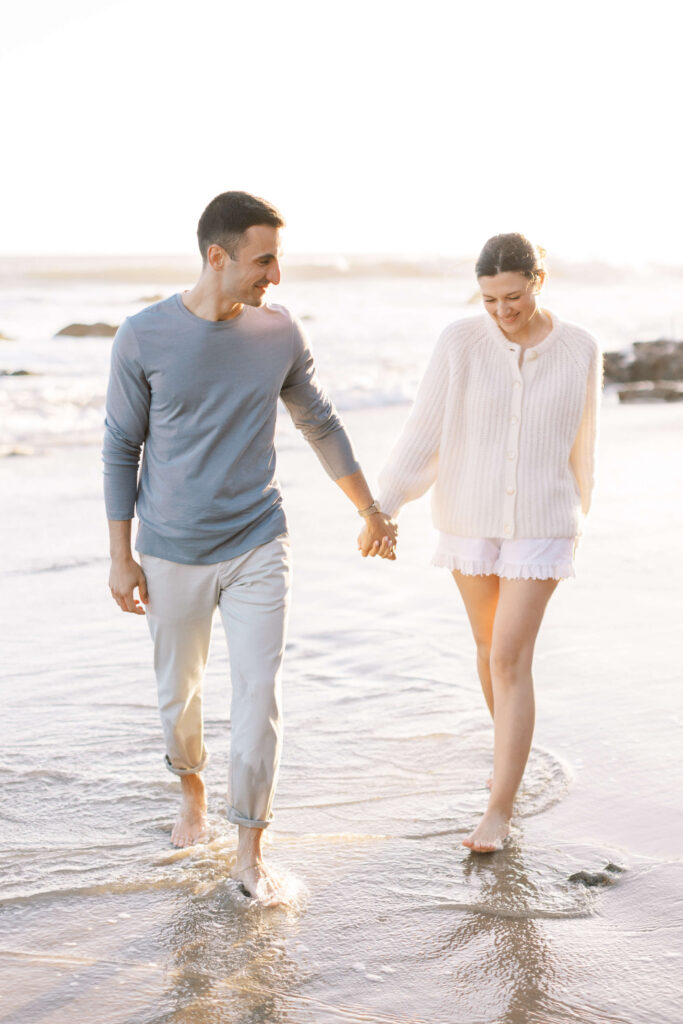 A smiling couple walks hand in hand along the shore, barefoot, with gentle waves at their feet. The man wears light pants and a blue shirt; the woman wears a white sweater and shorts. The scene is sunlit and relaxed. Copyright - Lisa Leanne Photography, California