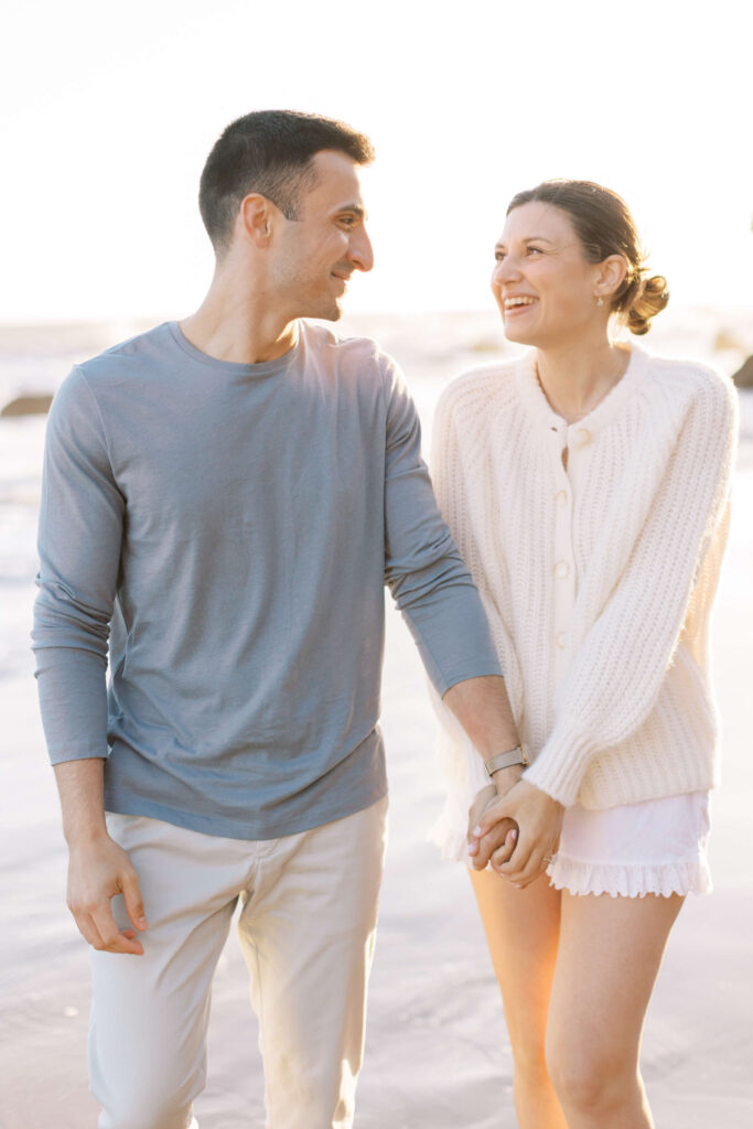 A smiling couple walks hand in hand along a beach at sunset, dressed in light casual clothing. They look at each other happily, with soft sunlight in the background and waves visible behind them. Copyright - Lisa Leanne Photography, California