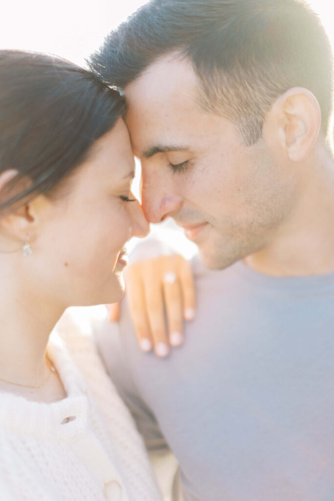 A couple stands close together with their foreheads touching, eyes closed and smiling softly. One person rests their hand on the other's shoulder, creating an intimate and tender moment in soft, natural light. Copyright - Lisa Leanne Photography, California