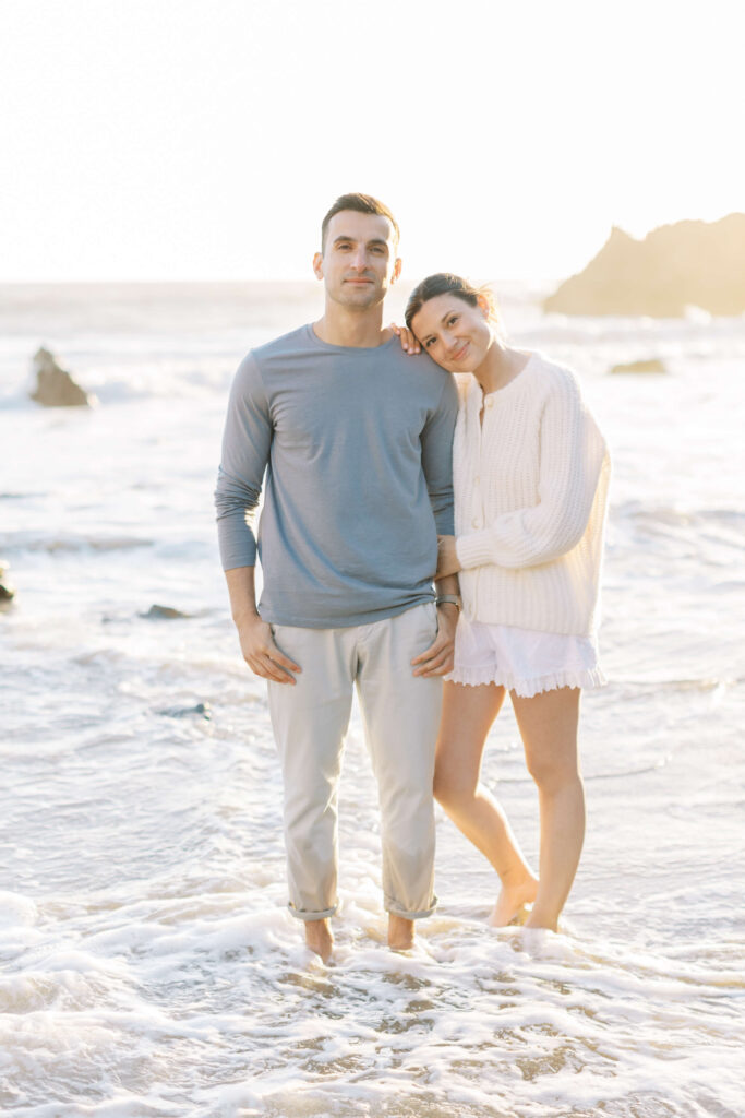 A couple stands in shallow ocean water at the beach. The woman, wearing a white sweater and skirt, leans her head on the man&rsquo;s shoulder. The man wears a light blue long-sleeve shirt and light pants. The scene is brightly lit. Copyright - Lisa Leanne Photography, California