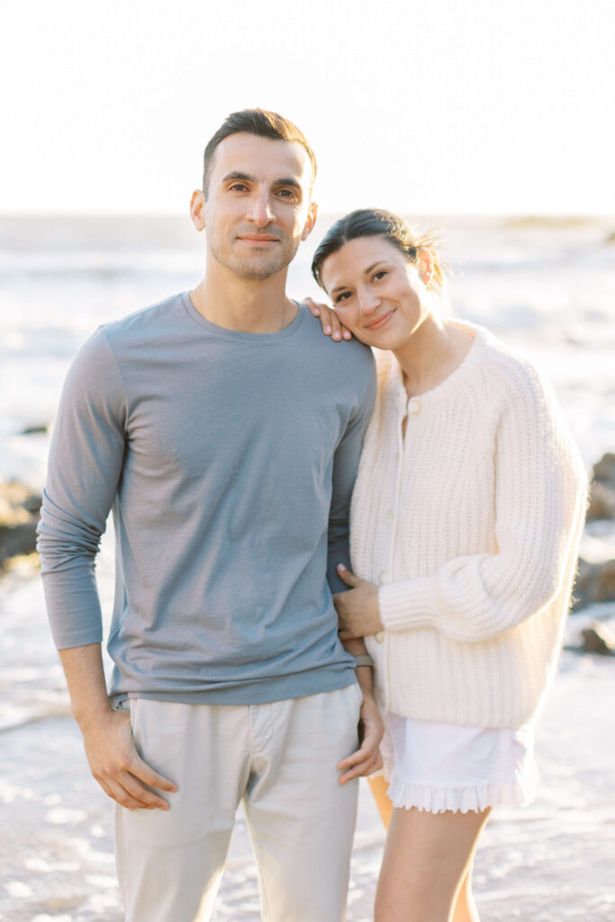 A couple stands on a beach; the woman leans her head on the man&rsquo;s shoulder, both smiling gently. He wears a light blue long-sleeve shirt, she wears a white sweater and shorts. The ocean and sand are in the background. Copyright - Lisa Leanne Photography, California
