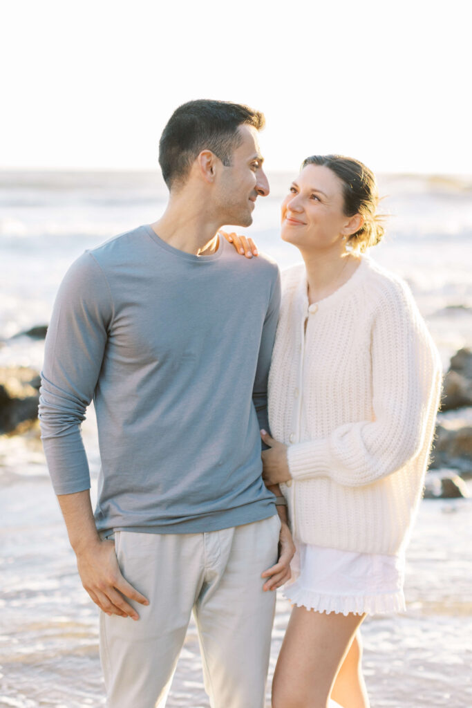 A couple stands on a beach. The man wears a light blue long-sleeve shirt and light pants, while the woman, smiling at him, wears a white sweater and shorts. They stand close together, with ocean waves in the background. Copyright - Lisa Leanne Photography, California