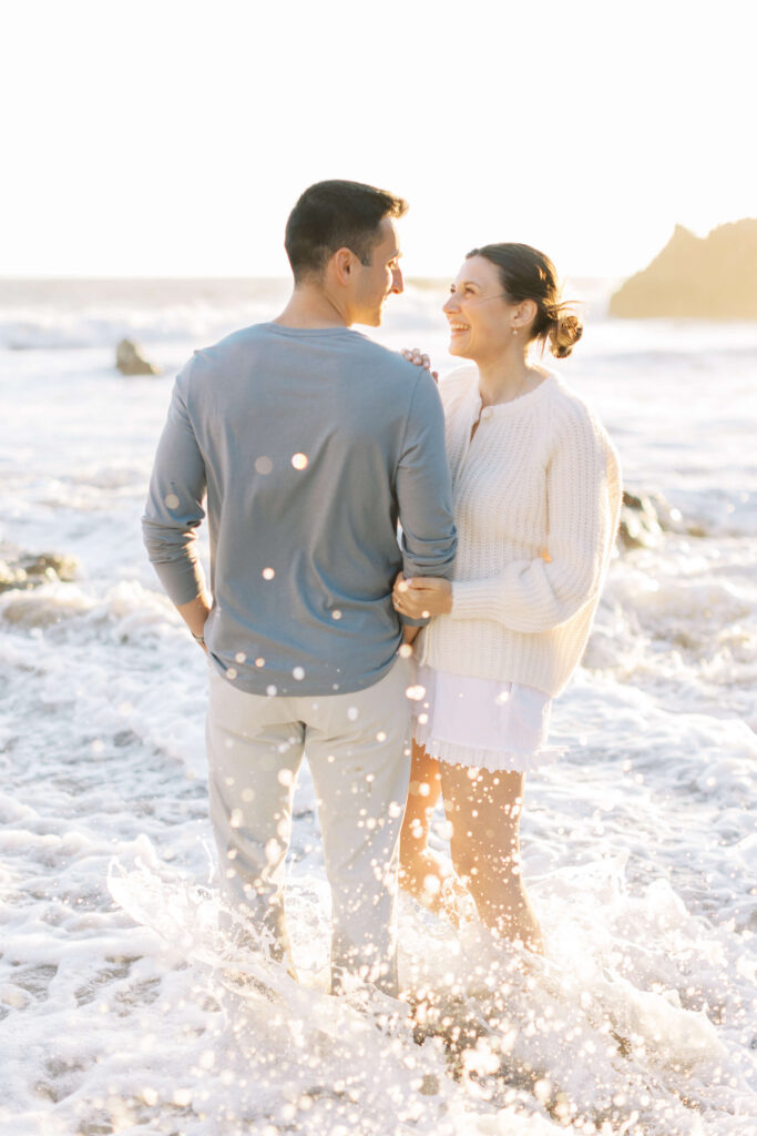 A couple stands in shallow ocean waves, smiling at each other as sunlight shines on them. Water splashes around their legs, and they appear joyful and relaxed by the beach. Copyright - Lisa Leanne Photography, California