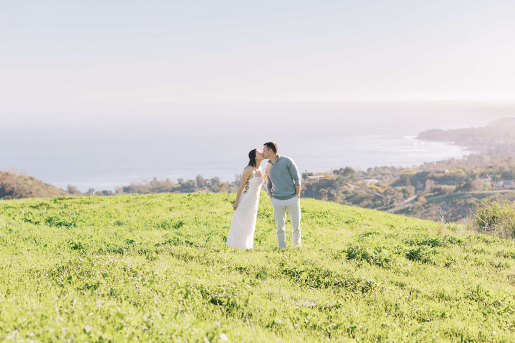 A couple stands on a grassy hill, kissing, with the ocean and a coastal landscape in the background under a clear sky. Copyright - Lisa Leanne Photography, California
