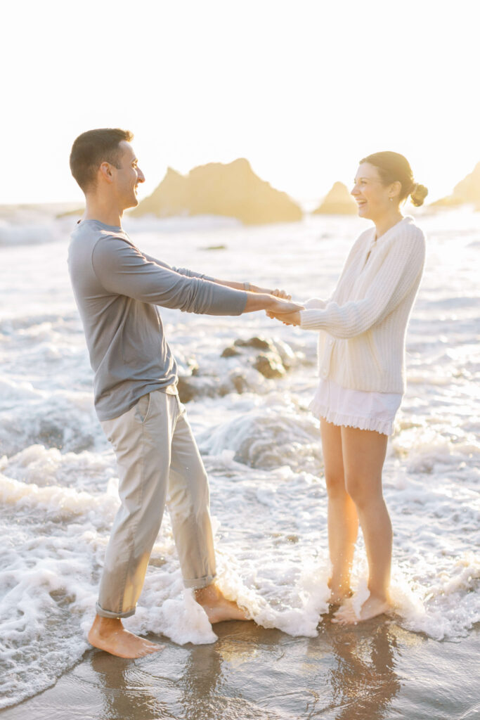 A man and woman stand barefoot in shallow ocean waves, holding hands and smiling at each other on a sunny beach, with rocks and bright sunlight in the background. Copyright - Lisa Leanne Photography, California