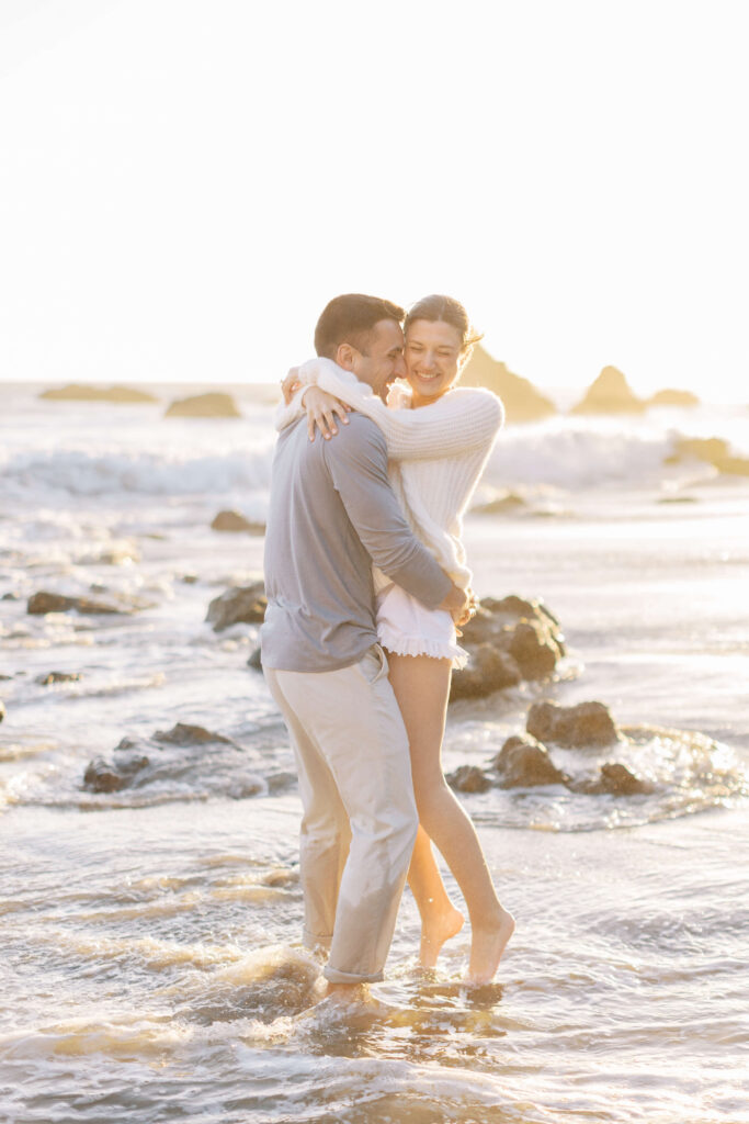 A couple stands in shallow ocean water at sunset, embracing and laughing. The woman wears a white sweater and shorts, while the man wears light-colored pants and a shirt. Waves and rocks are visible in the background. Copyright - Lisa Leanne Photography, California