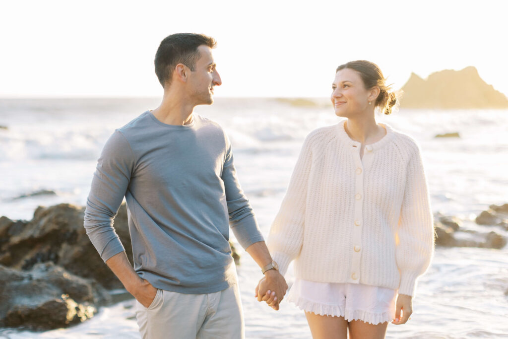 A couple stands on a rocky beach at sunset, holding hands and smiling at each other. The man wears a light blue shirt, and the woman wears a cream cardigan and white shorts. Waves and rocks are visible in the background. Copyright - Lisa Leanne Photography, California