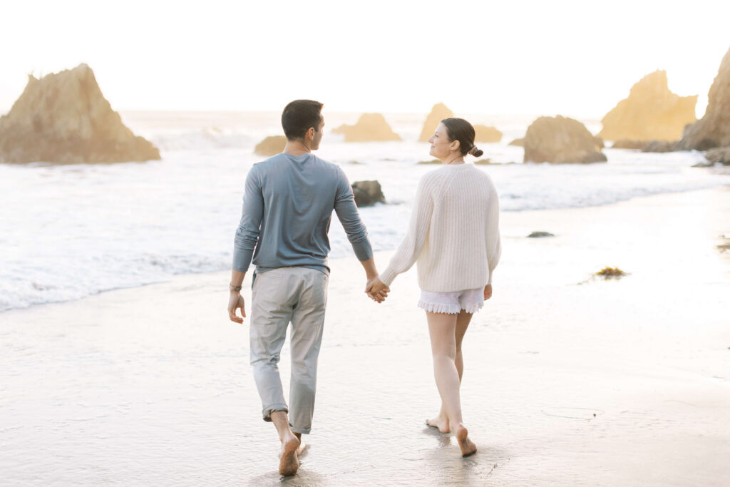 A couple walks barefoot, holding hands along a sandy beach with rock formations and gentle waves in the background at sunset. Copyright - Lisa Leanne Photography, California