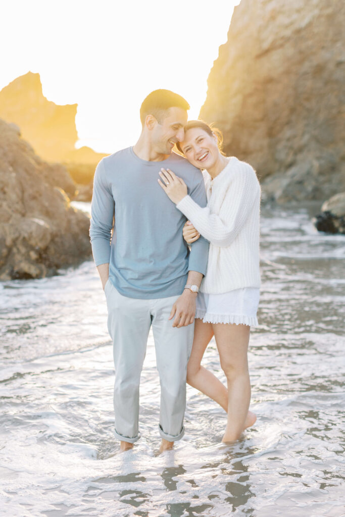 A smiling couple stands barefoot in shallow ocean water between rocks at sunset. The woman leans her head on the man's shoulder, both appear happy and relaxed. Copyright - Lisa Leanne Photography, California