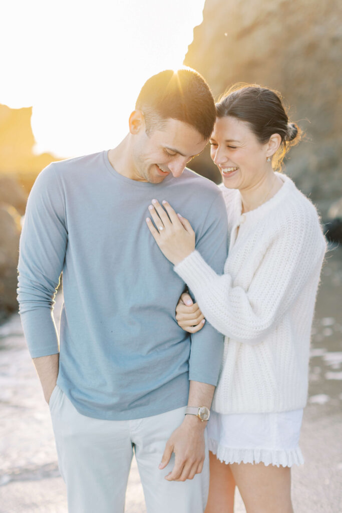 A smiling couple stands close together on a beach at sunset. The woman, wearing a white sweater, leans on the man's arm, showing an engagement ring, while the man in a light blue shirt looks down, both appearing happy. Copyright - Lisa Leanne Photography, California