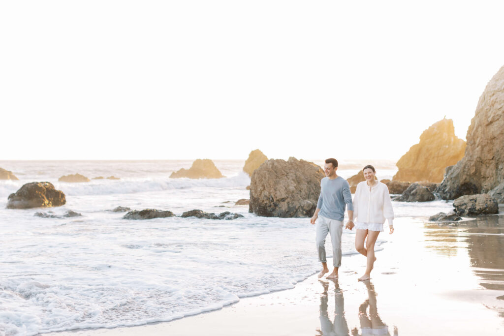A couple walks barefoot along a sandy beach near the ocean, holding hands and smiling. Rocks and gentle waves surround them under a bright, clear sky. Copyright - Lisa Leanne Photography, California