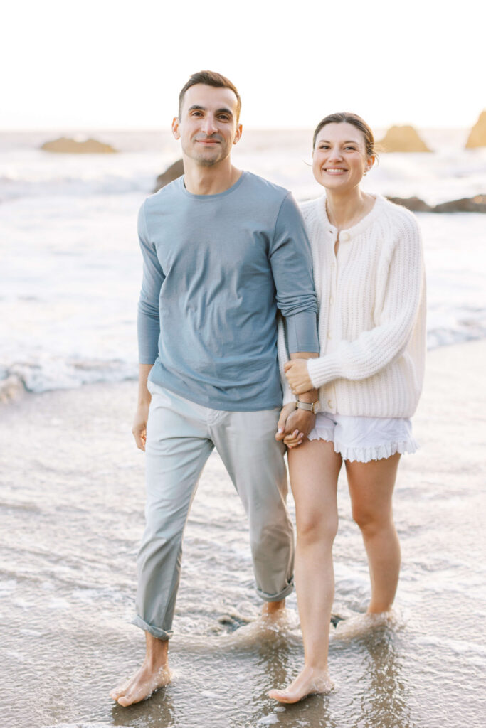 A smiling couple walks barefoot on the beach, holding hands. The man wears a light blue shirt and light pants, while the woman wears a white sweater and shorts. Gentle waves and rocks are visible in the background. Copyright - Lisa Leanne Photography, California